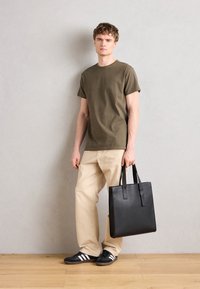 Svart lær tote-veske med to hanke, glatt tekstur og minimalistisk design. Modellen har på seg olivengrønn t-skjorte, beige bukser og mørke sko.