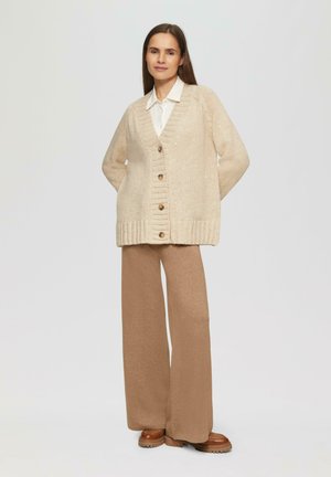 Beige gebreide cardigan met een V-hals, ribbelzoom en grote bruine knopen, gecombineerd met bruine wijde broek en bruine platformlaarzen.