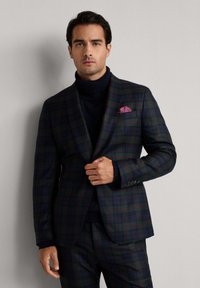 Uomo che indossa un completo scozzese verde scuro e blu navy con linee rosse, un maglione a collo alto blu navy e un fazzoletto da taschino a fantasia rossa, in piedi davanti a un muro chiaro.