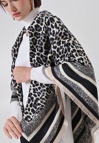 Écharpe en motif léopard avec des couleurs noir et crème, texture douce, bordures à franges et bords rayés contrastants. Portée sur un haut blanc.