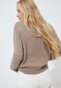 Femme blonde aux longs cheveux portant un pull marron et un pantalon blanc, légèrement de profil sur un fond blanc uni.