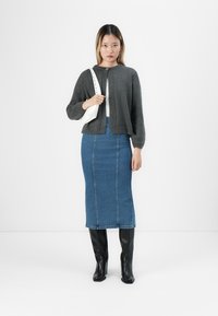 Žena v tmavě šedém cardigan, bílém topu, modré denimu midi sukni a černých vysokých botách, která stojí a nese bílou kabelku.