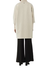 Beige oversized gebreid sweater met een hoge kraag, losse mouwen en een logodetail op de achterkant, gecombineerd met zwarte wijde broekspijpen.