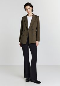 Junge Frau trägt ein olivfarbenes, doppelt geknöpftes Blazer, ein weißes T-Shirt, schwarze Schlaghose und schwarze Schuhe, während sie vor einem schlichten, hellen Hintergrund steht.