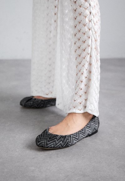 Black and gray patterned ballet flats with a zigzag design, featuring a rounded toe and a flat sole, worn with white textured fabric pants.