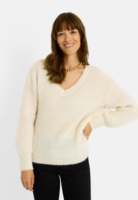 Jersey color crema hecho de material suave y texturizado, con escote en V, mangas anchas y ajuste relajado. Combina con pantalones negros.