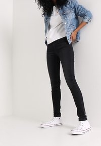 Chaqueta de denim, azul claro, con bolsillos frontales; sudadera gris; jeans ajustados negros; zapatillas blancas con detalles rojos. Tejidos texturizados, diseño casual.
