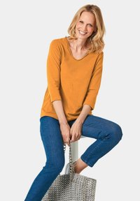 Senfgelbes V-Ausschnitt-Oberteil mit Dreiviertelärmeln, kombiniert mit blauen Skinny-Jeans. Eine silberne Studded-Handtasche wird seitlich gehalten.