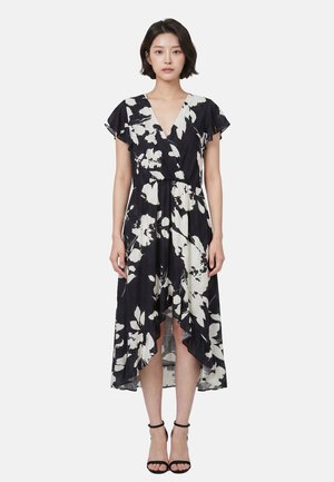 Donna in piedi che indossa un vestito a fiori bianco e nero con orlo alto-basso, maniche svolazzanti e tacchi neri con cinturino alla caviglia, su sfondo bianco semplice.