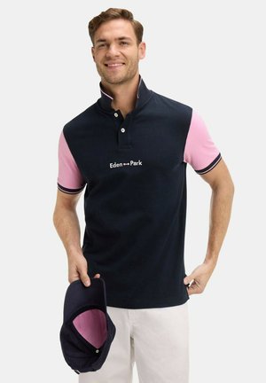 Un homme portant un polo bleu marine et rose, tenant une casquette assortie bleu marine avec une doublure rose, souriant devant un fond uni.