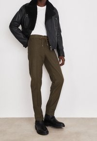 Svart läderjacka med fjärrkollare, vit t-shirt, olivgröna byxor med dragsko och svarta ankelstövlar på en neutral bakgrund.