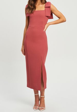 Femme portant une robe midi rose poudré sans manches à col carré avec des détails de nœuds sur les épaules et une fente avant, associée à des talons blancs à bride à la cheville.