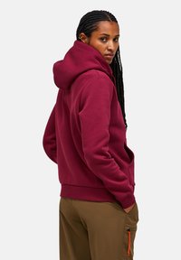 Felpa con cappuccio bordeaux dotata di tasca frontale, caratterizzata da una texture morbida e polsini a costine. Indossata con pantaloni verde oliva e un dettaglio arancione sulla zip.