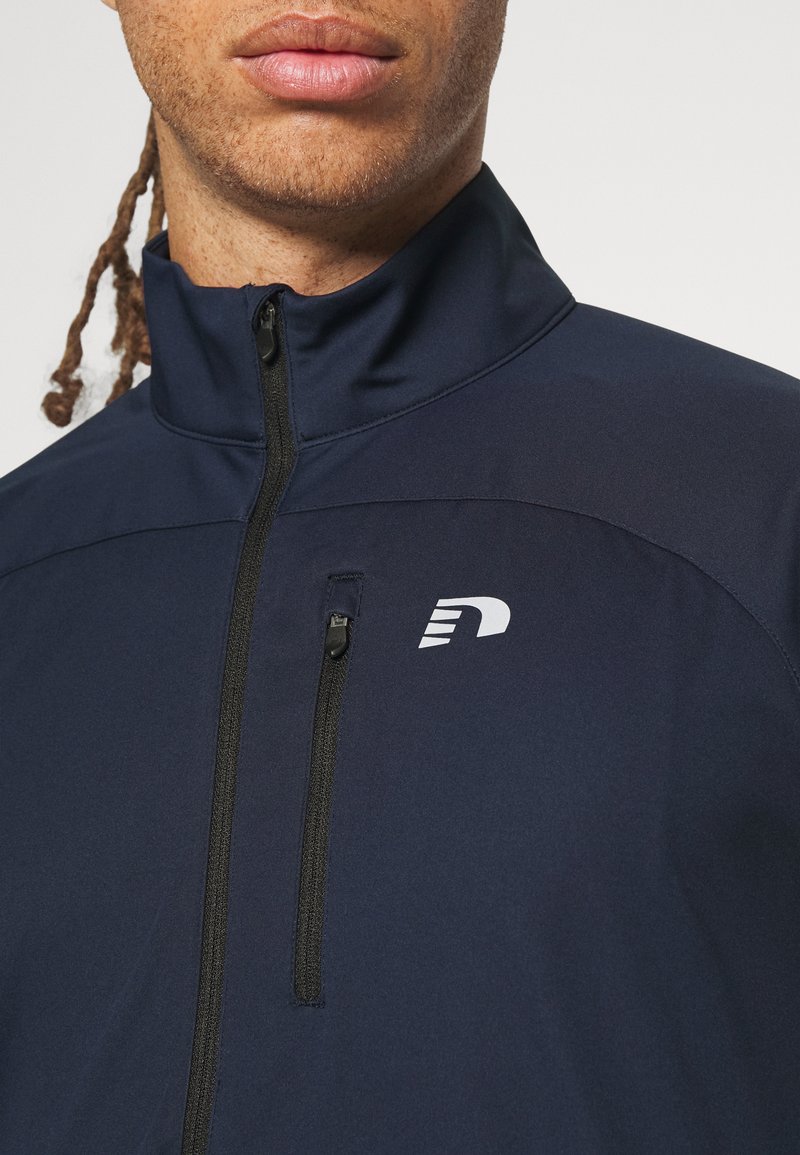 Marineblaue Zip-Jacke mit hohem Kragen, ausgestattet mit einem kleinen weißen Logo auf der Brust und einer Reißverschlusstasche. Glattes, leichtes Material.