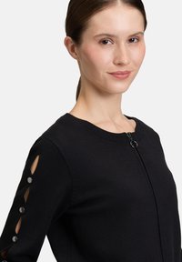 Pull noir à fermeture éclair avec un col rond. Présente des découpes sur les manches accentuées par des boutons noirs. Fabriqué en un tissu tricoté lisse.