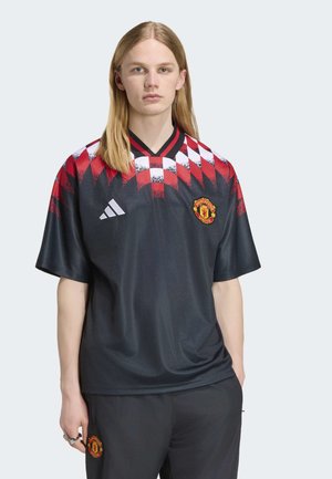 Ung person med langt blondt hår iført sort Manchester United fodboldtrøje med rødt og hvidt ternet mønster på skuldrene.
