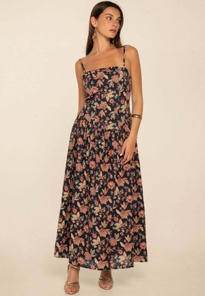Mujer con vestido negro sin mangas hasta el tobillo con estampado floral rosa y beige, pulsera de oro, pendientes y sandalias doradas abiertas en los dedos.