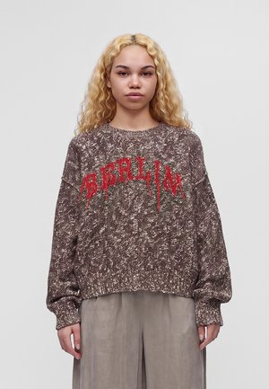 Jeune personne aux longs cheveux blonds bouclés porte un pull marron et blanc tacheté avec un texte rouge et un pantalon gris ample sur un fond uni.