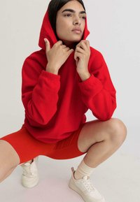 Felpa rossa con cappuccio, abbinata a pantaloni corti da ciclismo rossi. Il modello indossa scarpe da ginnastica bianche e calzini crema, seduto in posizione accovacciata.