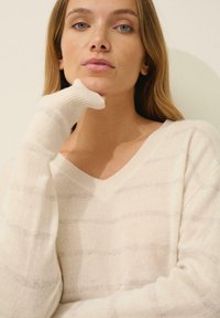 Sweter kremowy z dekoltem w serek i długimi rękawami, charakteryzujący się subtelnymi poziomymi paskami i miękką teksturą. Model pozuje z ręką przy brodzie.