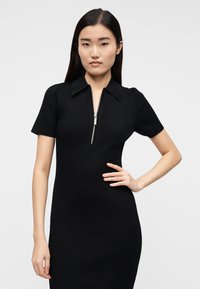 Črna rebrasta obleka s kratkimi rokavi, z zadrgo spredaj in ovratnik kakršne vrste. Oblikovana, da tesno prilega, z gladko teksturo.