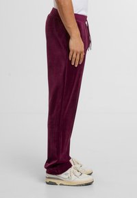 Person in tiefvioletten Velours-Sweatpants und weißen Sneakers, seitlich vor einem schlichten hellen Hintergrund stehend.