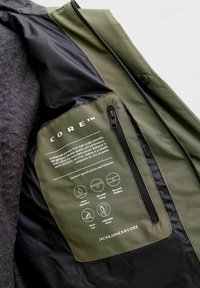 Interiörficka i olivgrön jacka med tryckta plaggspecifikationer, svart dragkedja och ikoner för vattenavvisande, återvunnen foder, vaddering, stormmudden och justerbar huva.