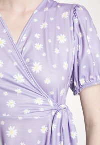 Vestido envolvente roxo feito de tecido suave, com margaridas brancas. Mangas bufantes e um laço na cintura; textura suave e leve.
