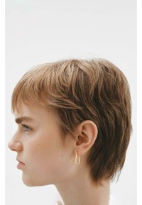 Pendiente de aro de metal dorado con forma rectangular, llevado en la oreja izquierda. El cabello corto y texturizado enmarca la cara, contra un fondo neutro.