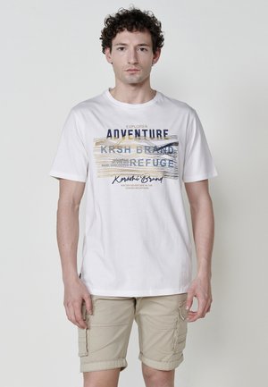 T-shirt bianca in cotone con grafica colorata e linee, abbinata a pantaloni cargo beige, con tasche e orlo arrotolato.
