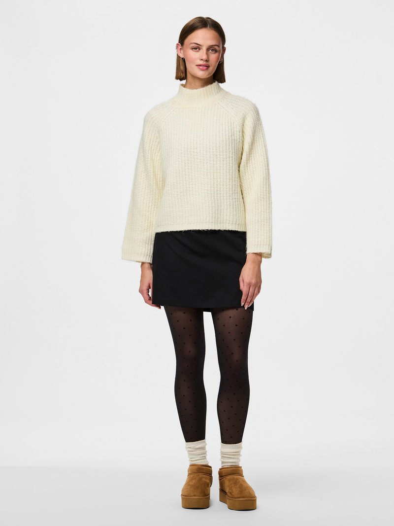 Femme debout portant un pull en maille crème, une mini-jupe noire, des collants noirs à pois, des chaussettes crème et des chaussures à plateforme marron.