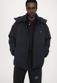 Puffer jacket in dark navy with a grid pattern, featuring a hood, zip closure, and front pockets. Worn over a black polo shirt with a grid design.