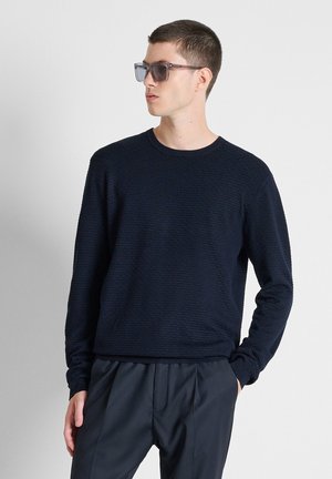 Man draagt een donkere gestructureerde trui, donkere broek en zonnebril, kijkt opzij met één hand in de zak tegen een effen achtergrond.