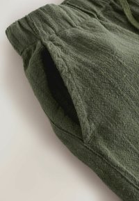 Pantalon en tissu texturé vert foncé avec une ceinture élastique et une poche latérale visible sur un fond clair.