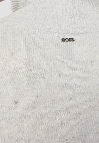 Helle graue Strickjacke mit hohem Kragen, die eine dezente Textur aufweist und ein kleines schwarzes "BOSS"-Logo auf dem Rücken hat.