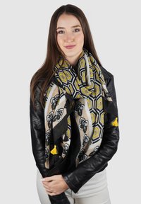 Zwarte en gele sjaal met een bloemendessin en franjes, gedrapeerd over een zwart leren jas. Lichte grijze achtergrond.