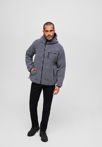 Chaqueta de forro polar gris con capucha, frente con cremallera y dos bolsillos laterales. Presenta un tejido texturado y un bolsillo en el pecho con cremallera.