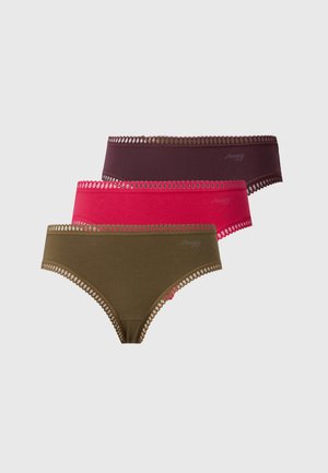 Kvinde med langt mørkt hår iført en beige bh og rødt undertøj, stående barfodet mod en ensfarvet hvid baggrund.