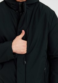 Zwarte jacket heeft een ritssluiting aan de voorkant en een capuchon. Het materiaal lijkt glad en geïsoleerd, met een zichtbare zijzak. Volledige rits en trekbanden.