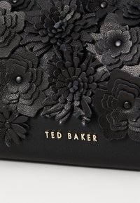 Crna kožna površina s višeslojnim cvjetnim aplikacijama različitih tekstura. Sadrži zlatni logo "TED BAKER" na dnu.