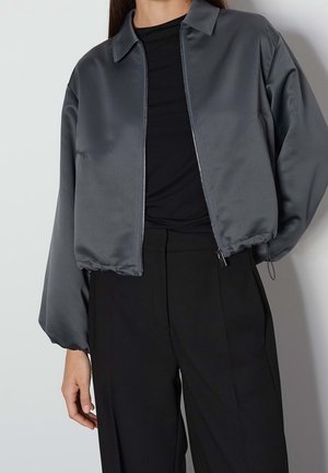 Nainen pukeutuneena tummanharmaaseen lyhennettyyn vetoketjutakkiin, jossa on kaulus, mustan paidan päällä ja mustat korkeavyötäröiset housut yksinkertaisen taustan edessä.