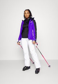 Lila und schwarze Skijacke, schwarzes Baselayer, weiße Hose und schwarze Skischuhe. Hält pinke Skistöcke mit einem eleganten, sportlichen Design.