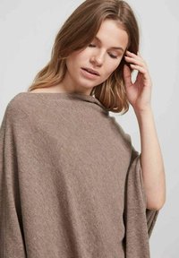 Ruskea neuleponcho, jossa on väljä malli, pehmeä rakenne, leveät hihat ja kauloseinämuotoilu, joka laskeutuu helposti olkapäiden yli.