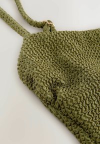 Tissu texturé vert avec un design smocké. Présente des bretelles réglables et un accent en matériel doré.