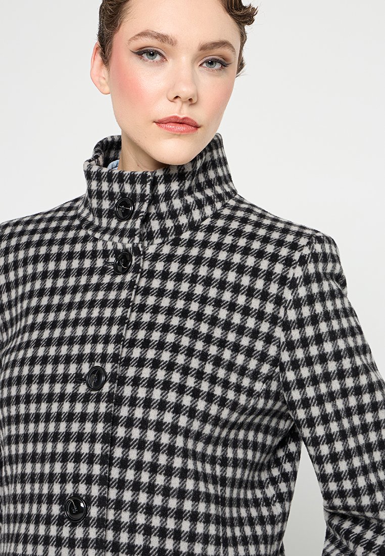 Zwart-wit houndstooth jas met een hoge kraag, een slim fit ontwerp en vijf zwarte knopen aan de voorkant. Zacht, gestructureerd materiaal.