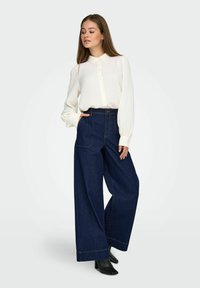 Chemisier blanc avec un col texturé, associé à un pantalon en denim sombre à jambes larges avec des coutures contrastées et des poches latérales. Bottines noires.