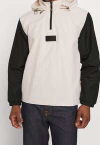 Anorakjacka med vit kropp och svarta ärmar, med en front halvdragkedja, justerbar huva och en liten svart logotyp på bröstet.