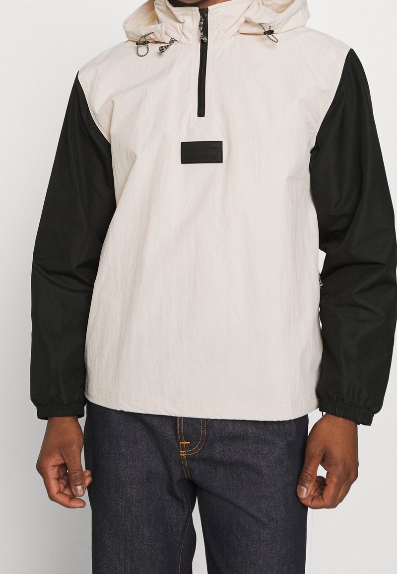 Anorakjacka med vit kropp och svarta ärmar, med en front halvdragkedja, justerbar huva och en liten svart logotyp på bröstet.