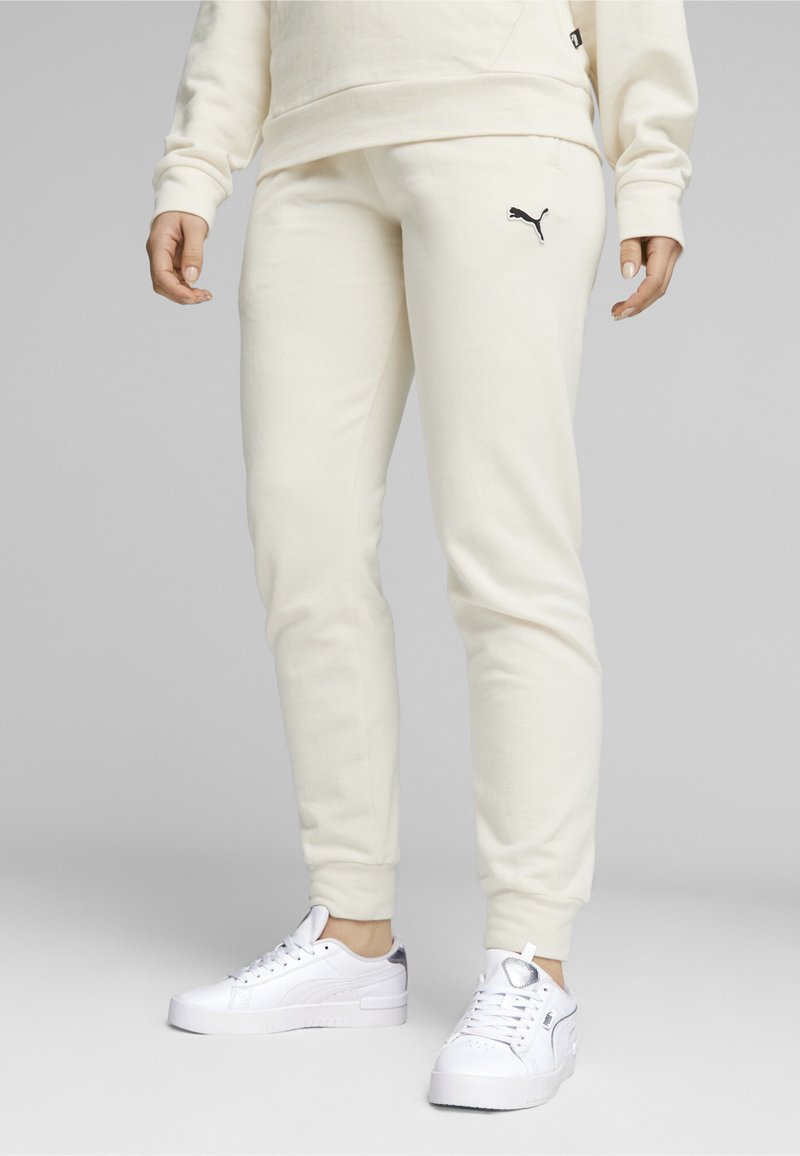 Pantalones de chándal color crema hechos de una tela suave, con un diseño ajustado y un logo negro de Puma en el lado izquierdo. Zapatillas blancas completan el look.