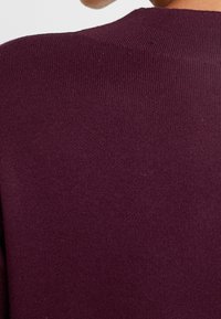 Burgunderfarbener Strickpullover mit gerippter Struktur und Rundhalsausschnitt, der eine glatte Oberfläche und einen leichten Glanz im Stoff aufweist.